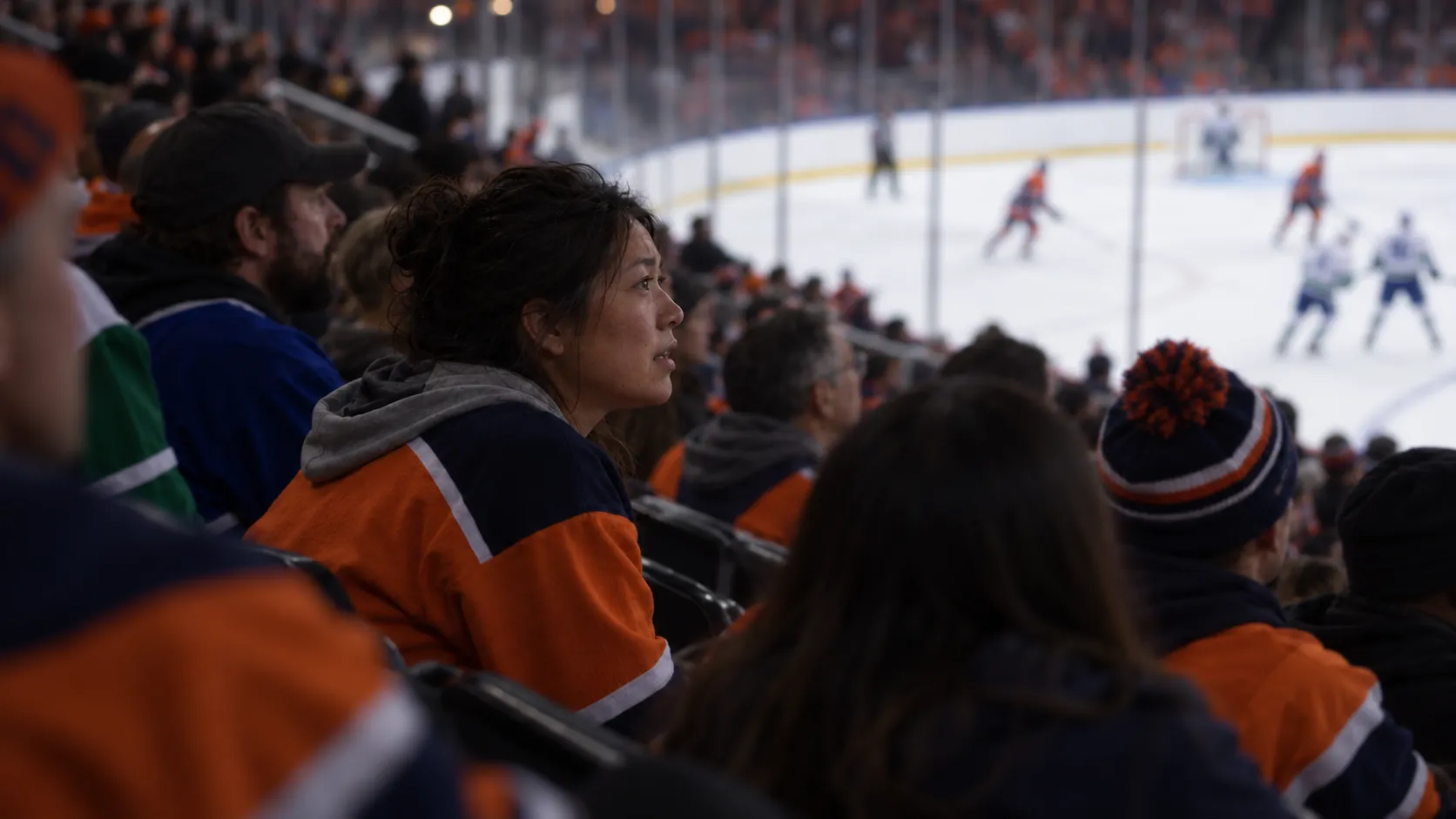 Hockey fans in Canucks blue and green and Oilers orange and navy watch from dimly lit stands  with the ice softly blurred beyond
