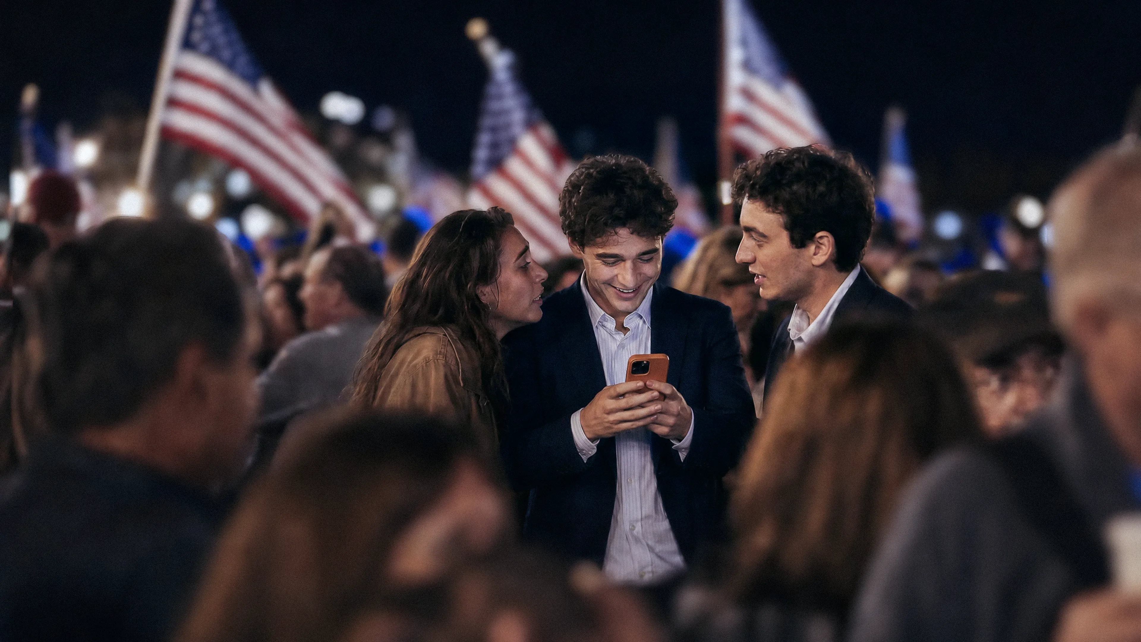 3 people looking at a phone on a political rally with American flags in the background
