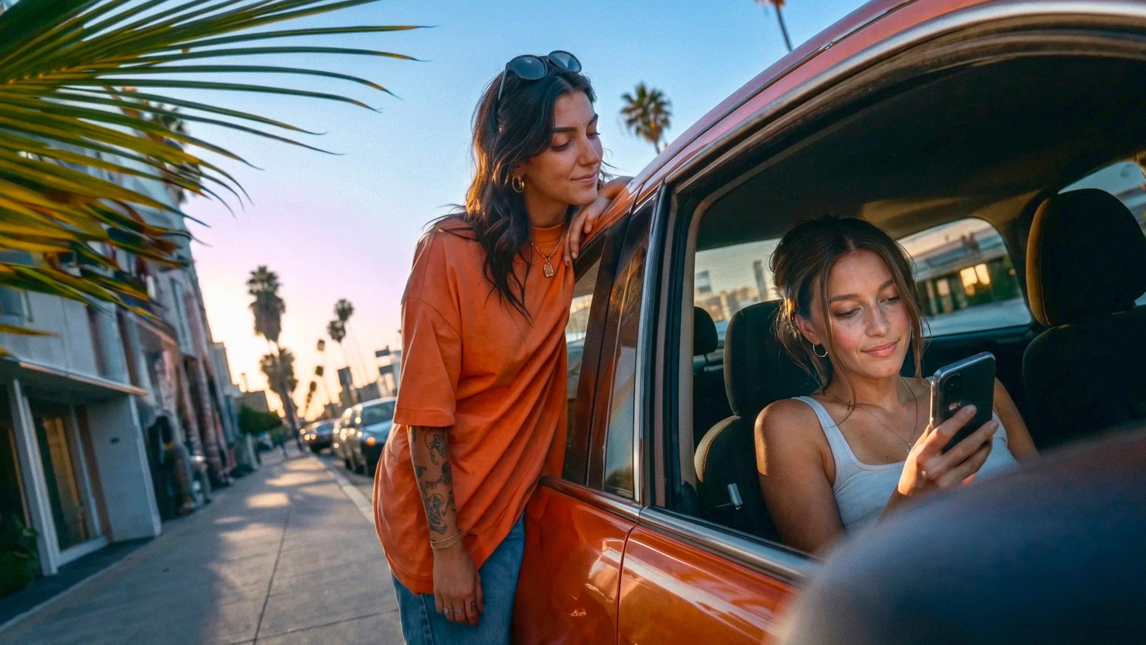 two women looking at a phone one is standing outside with a palm tree in the background and the other one is in the car