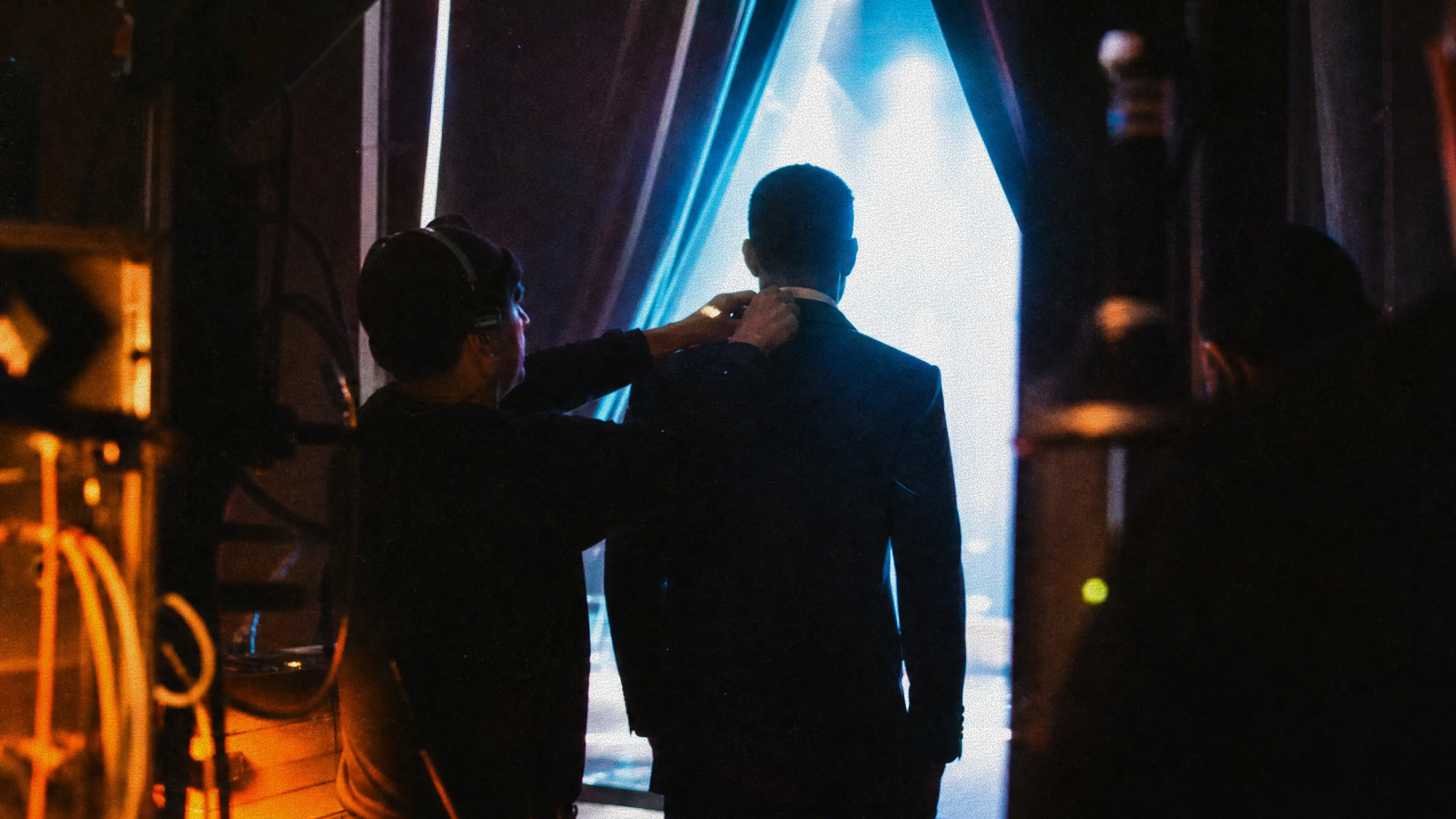 A crew member adjusts the collar of a man in a dark suit backstage as he faces a brightly lit blue entrance moments before stepping on stage